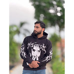 Bearded man in black graphic hoodie and jeans standing outdoors with blurred green trees background