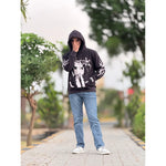 Man in black graphic hoodie, blue jeans, and sneakers standing on a paved path with greenery