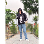 Man in black graphic hoodie, blue jeans, and sneakers standing on a paved path with greenery
