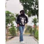 Man in black graphic hoodie and blue jeans standing on a paved path with greenery outdoors