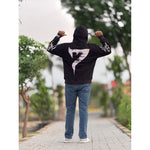 Person wearing black hoodie with large white number 7 design, blue jeans, standing on paved path outdoors