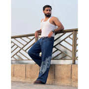 Bearded man wearing white tank top and blue jeans with dragon print leaning on railing outdoors