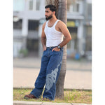 Man in white tank top and blue jeans with dragon print leaning on tree outdoors